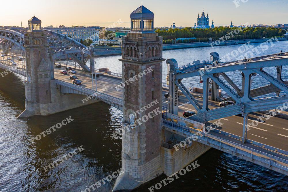 На снимке запечатлен Большеохтинский мост, перекинутый через реку Неву, с панорамным видом на Смольный собор. Массивные опоры моста подчеркивают его инженерное величие, а открывающийся на заднем плане Смольный собор добавляет композиции элегантности. Вода Невы и вечернее освещение придают кадру особую атмосферу спокойствия и гармонии. Это сочетание современности и исторического наследия символизирует уникальный облик Петербурга.